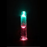 The RealRock Wireless Rechargeable LED Vibrating 6" by Shots America, a transparent cylindrical dildo, glows with pink and teal lights from below, creating a vibrant effect in the dark.